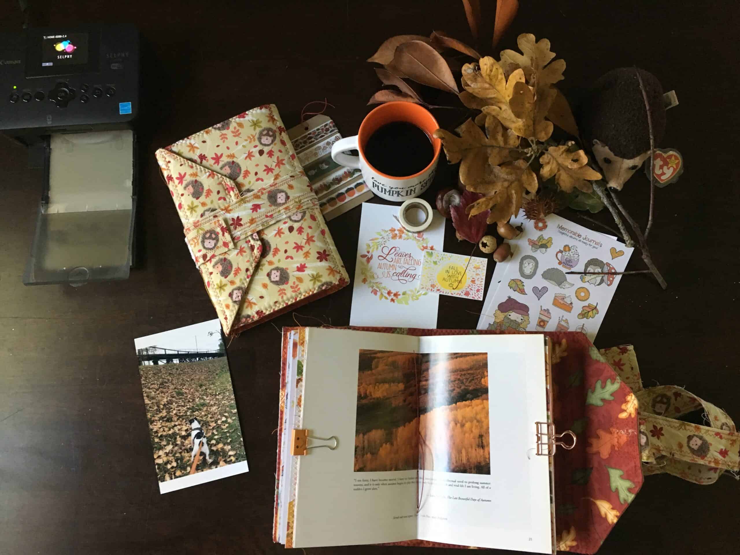 autumn themed journal desktop scene showing a closed journal covered in a hedgehog fabric and an open journal showing an autumn scene. Journals are surrounded by a photograph of walking a dog in the autumn leaves, an orange mug filled with coffee and journaling accessories related to hedgehogs in autumn theme.